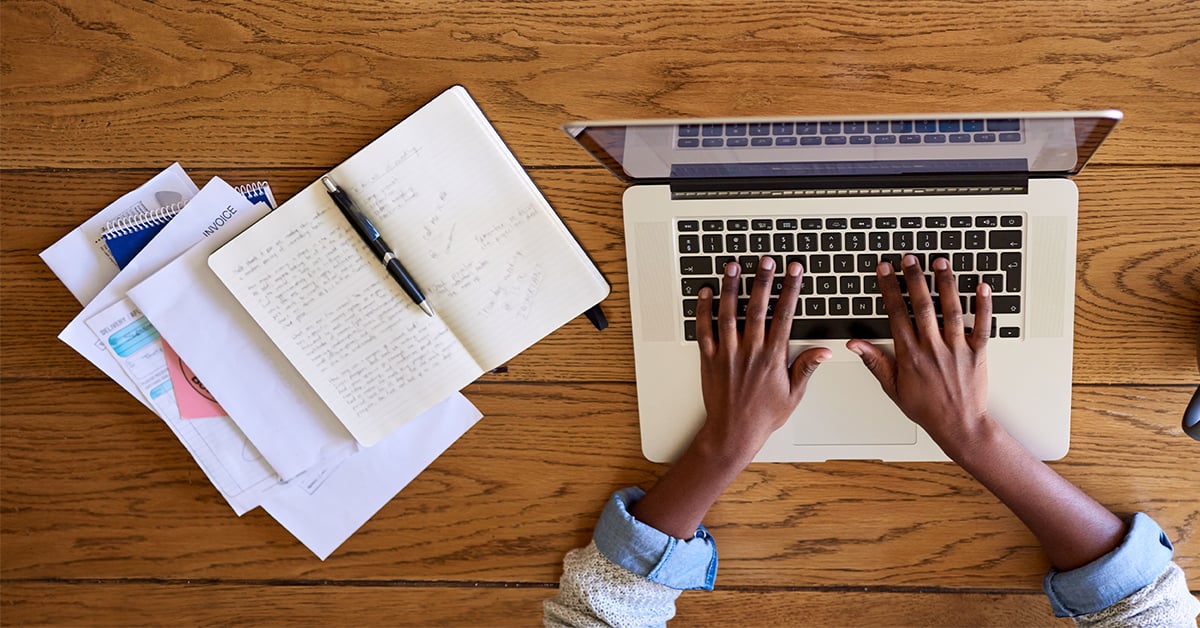 A woman's hands typing her clinical research paper beside a notebook and papers. 