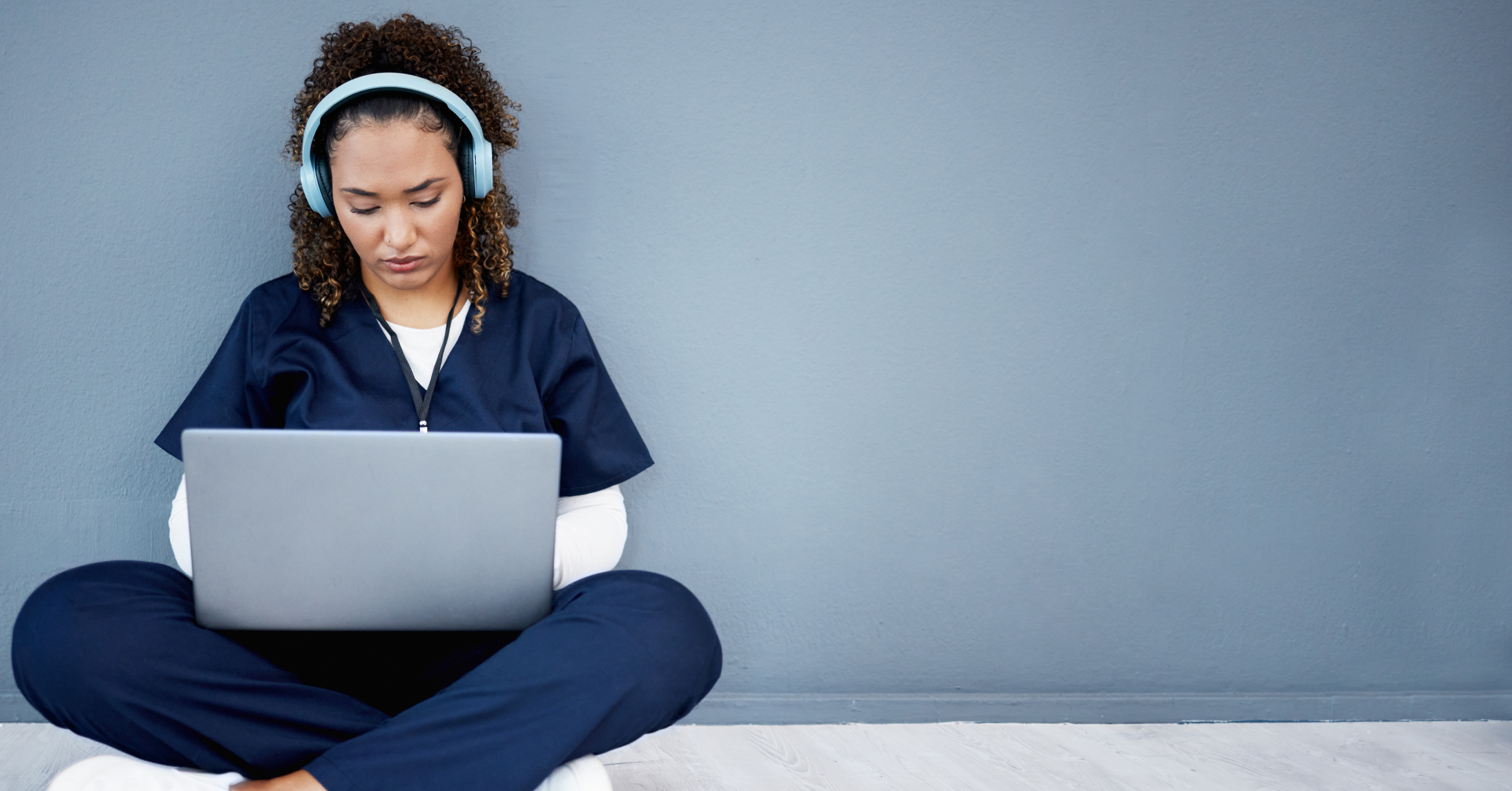 A young female physician wearing scrubs and headphones looking at a laptop: the best medical podcasts.