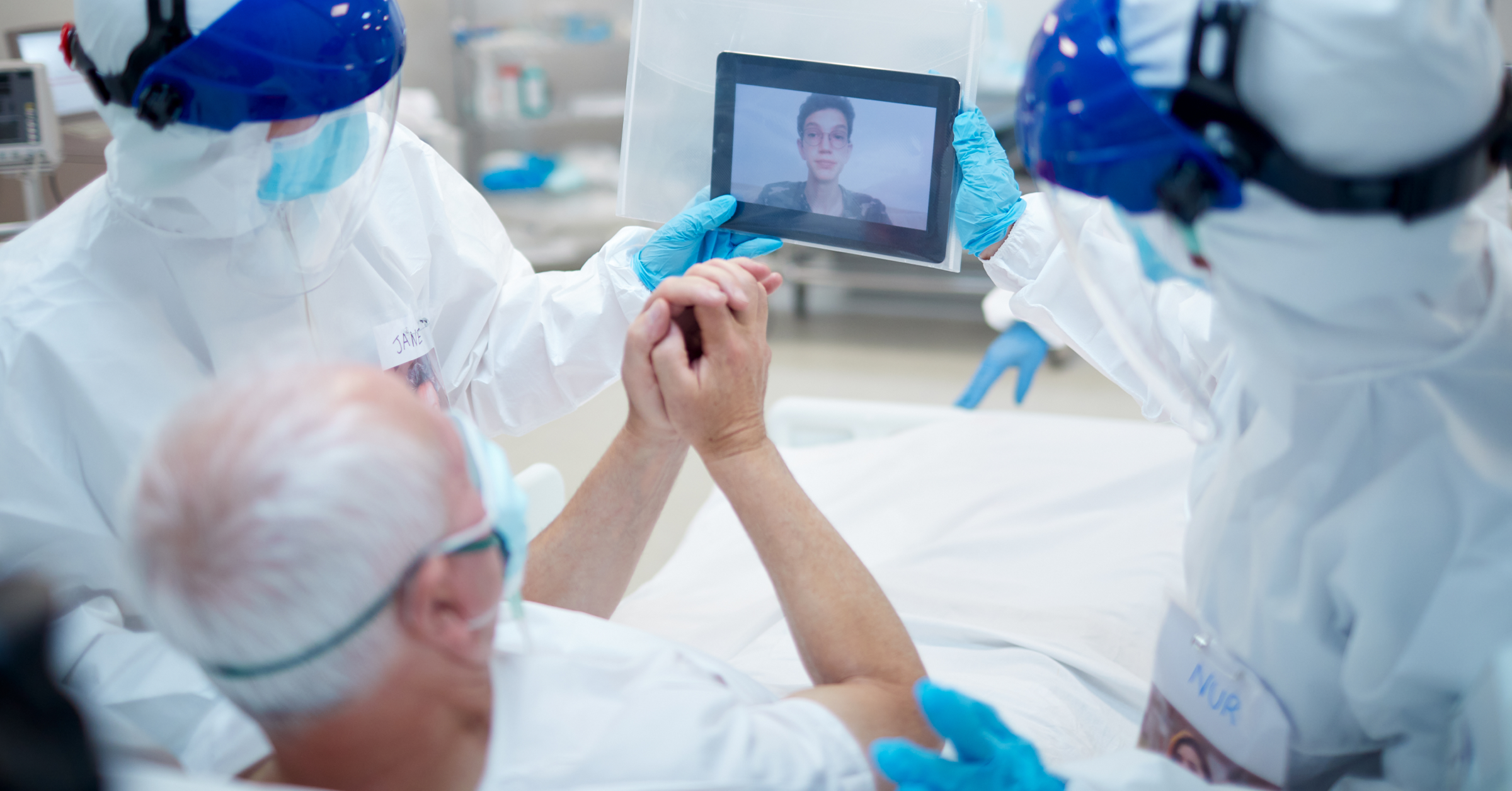 Zwei Fachkräfte in Schutzkleidung halten ein Tablet, damit ein Patient mit einer nahestehenden Person ein Videotelefonat führen kann.