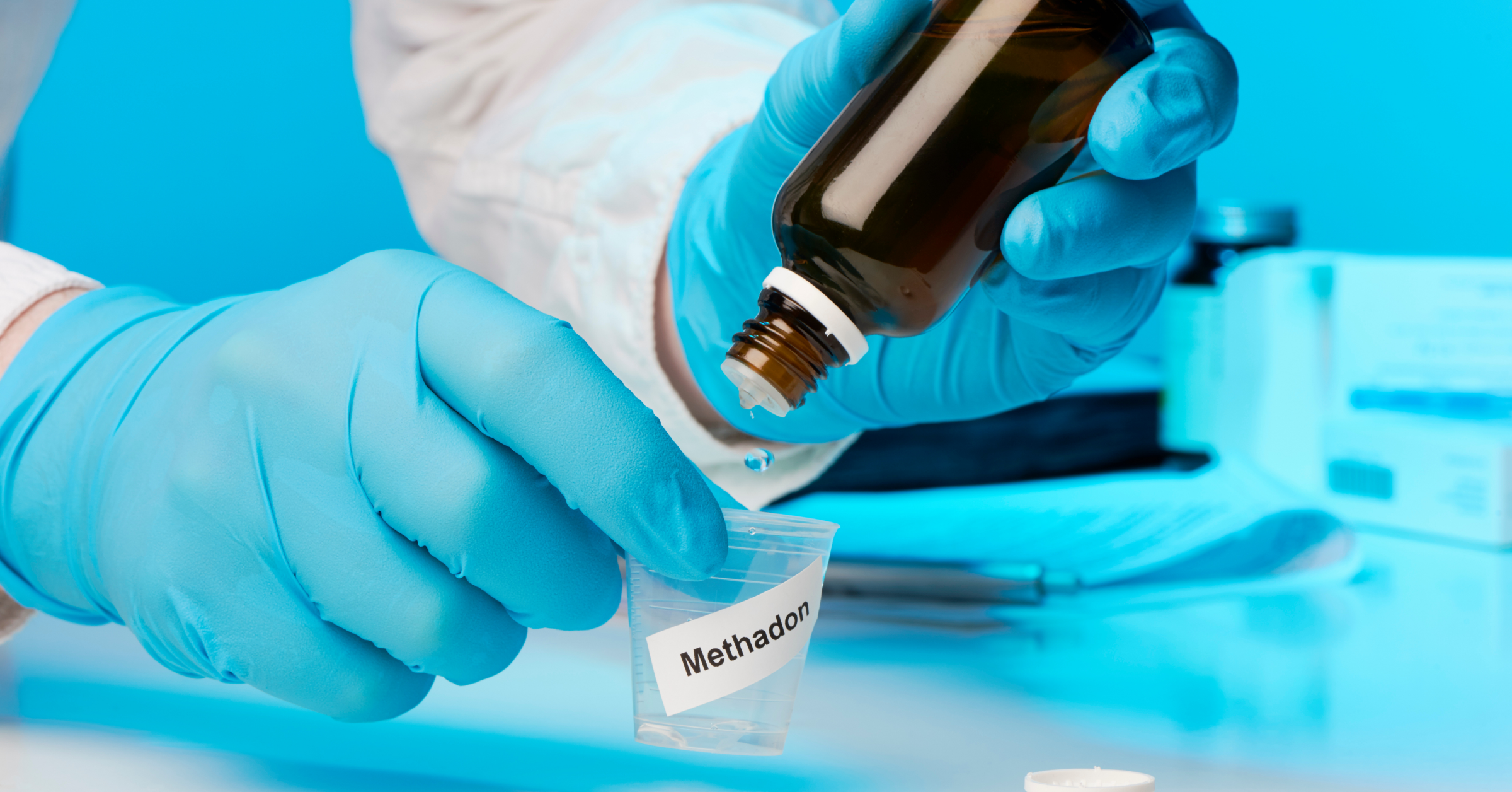 Eine Hand in Gummihandschuhen träufelt Methadon in einen Medikamentenbecher. Opioid-Substitution im AMBOSS-Blog