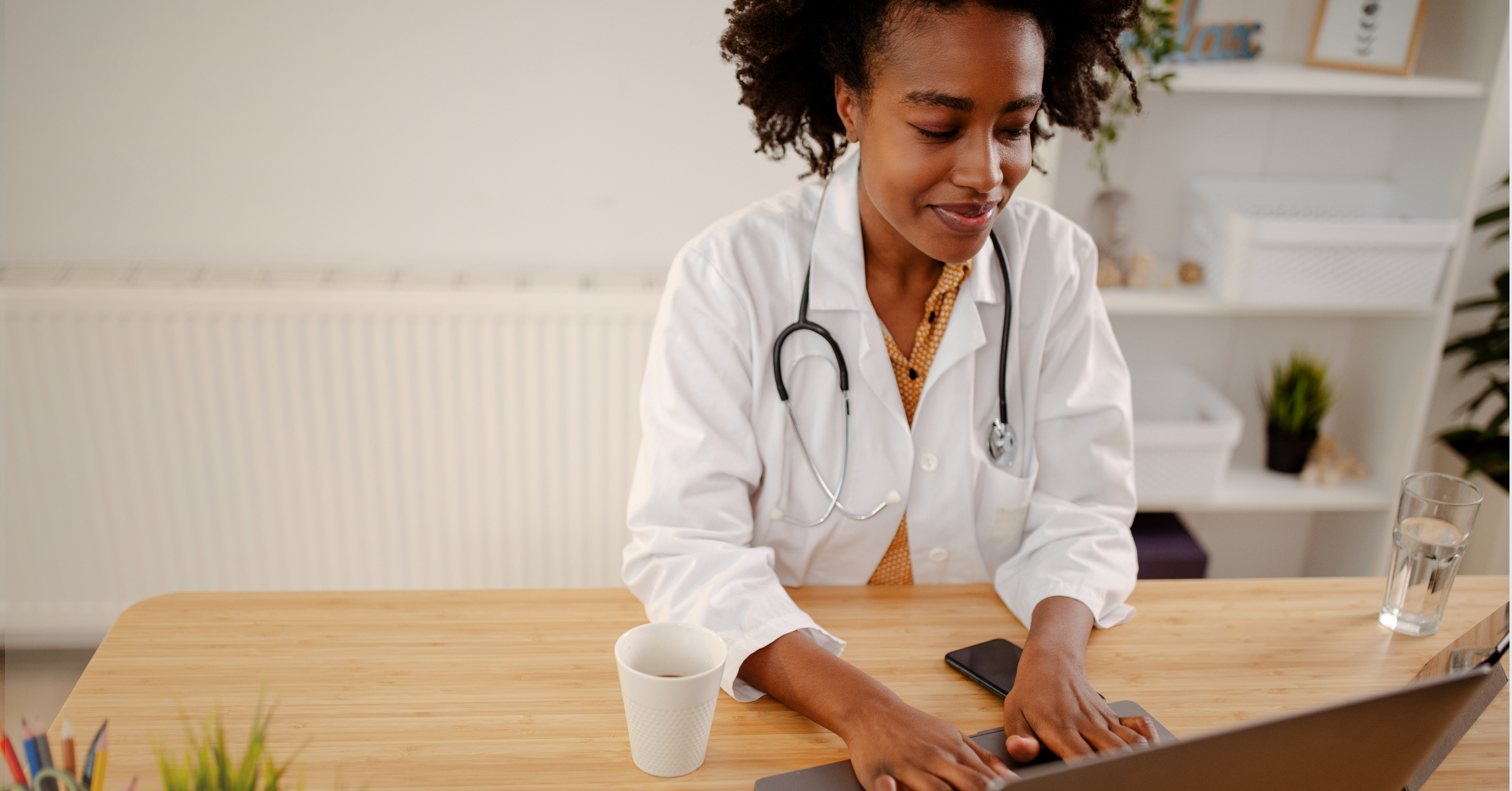 A doctor in a white coat and stethoscope types on a laptop. 