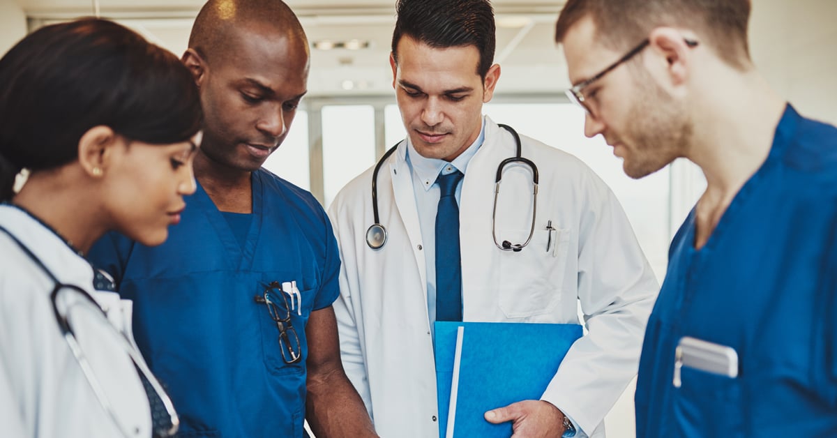 Medical students in blue scrubs and white coats huddle together during one of their third year clerkships.