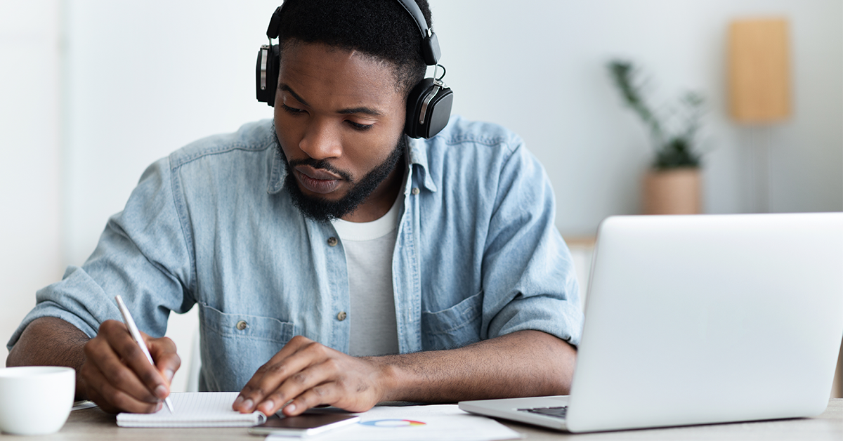 A man wearing headphones studies with a notepad and laptop.
