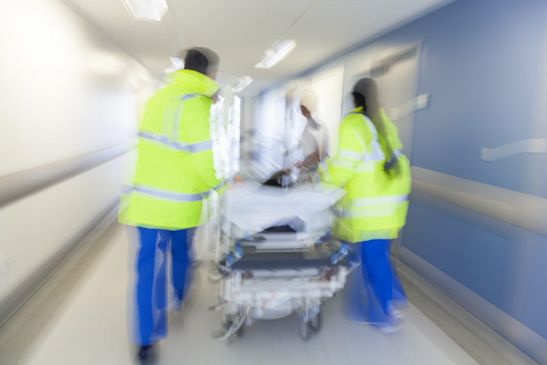 Zwei Sanitäter schieben eine Person auf der Patientenliege durch den Gang eines Krankenhauses.