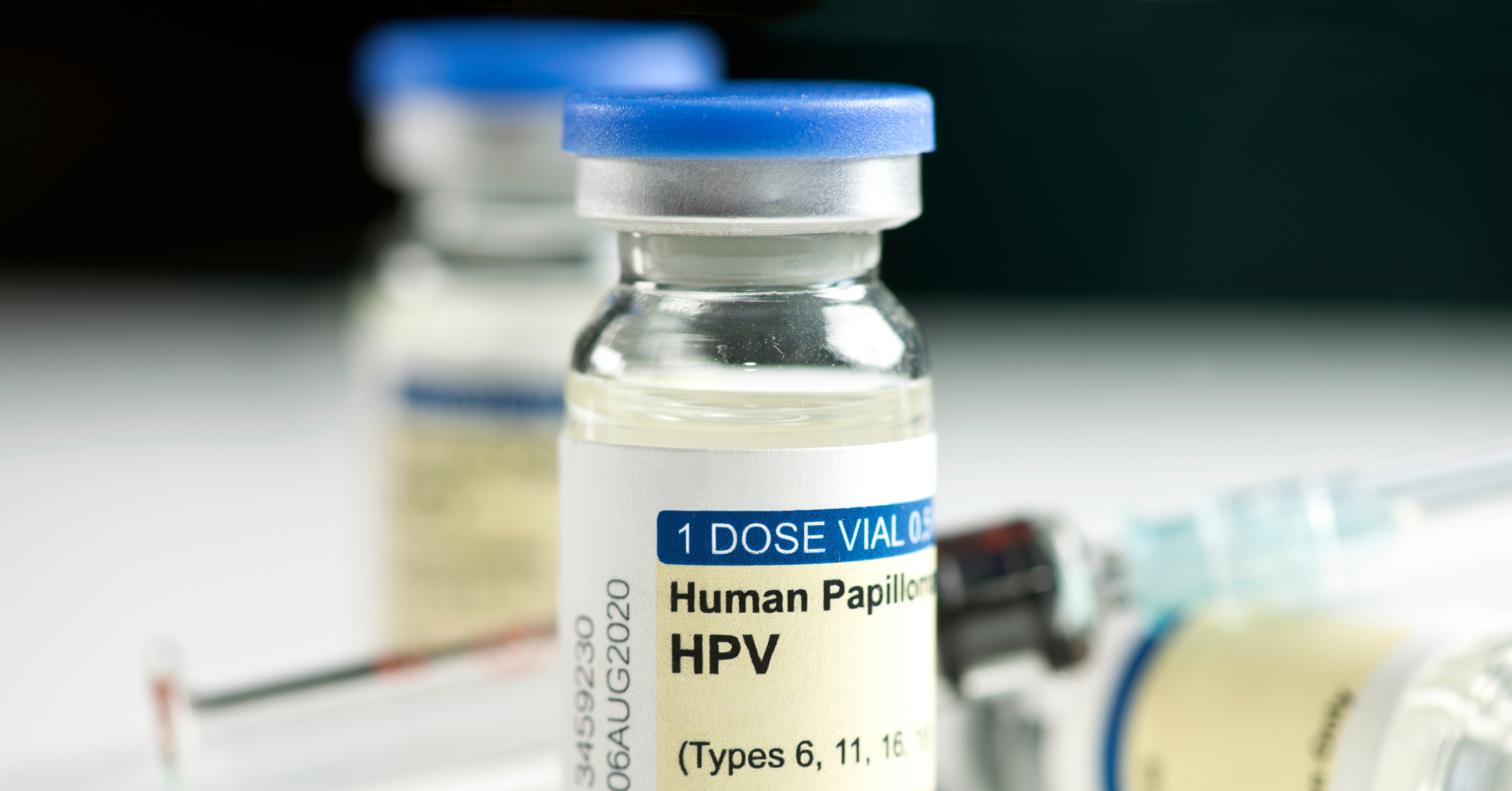Ein Glasbehältnis mit einer Dosis HPV-Impfstoff steht vor einer Spritze. HPV im AMBOSS-Blog.